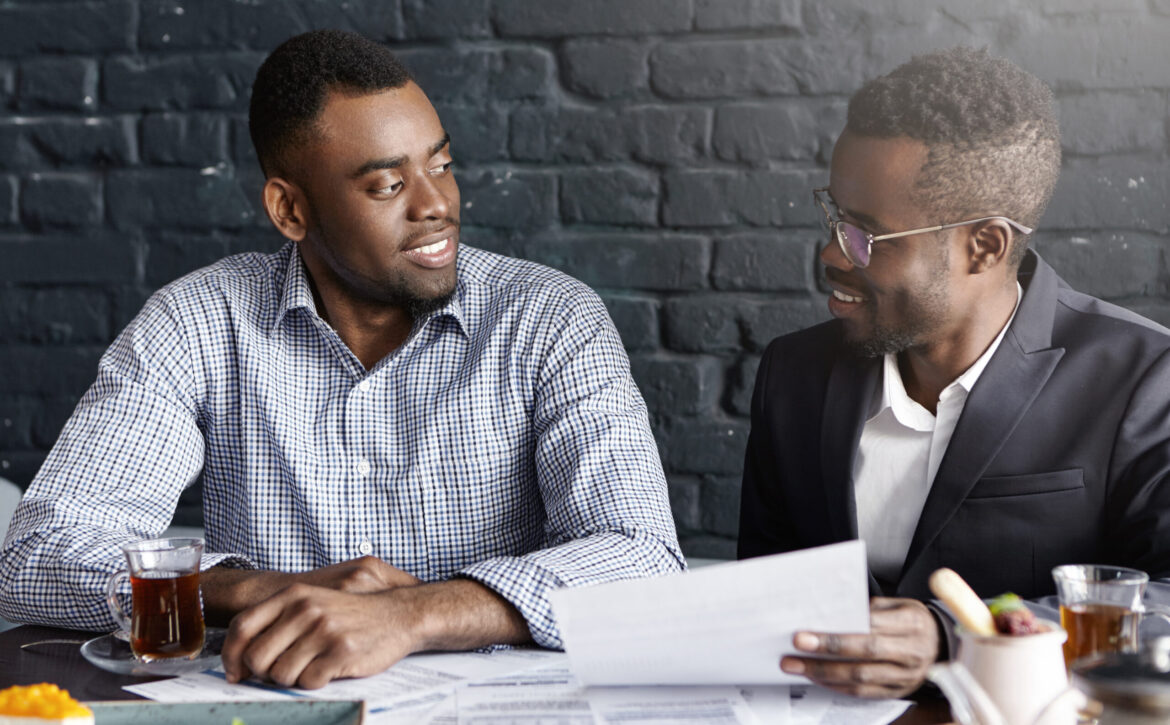 Two confident and successful dark-skinned business partners having nice conversation, discussing plans, sharing ideas and vision, sitting at table with papers during lunch break at modern restaurant